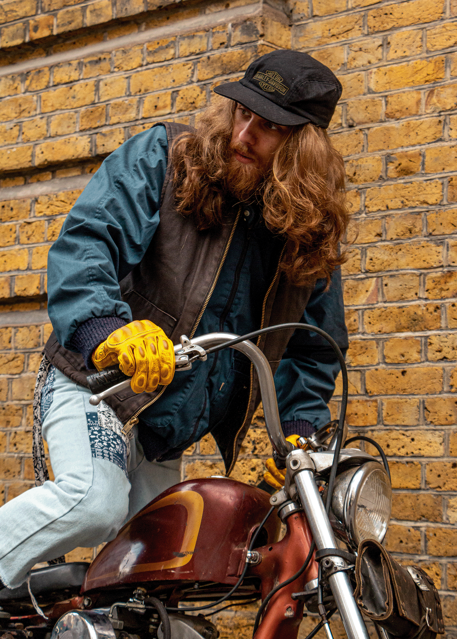 Person with long hair and beard sitting on a motorcycle against a brick wall