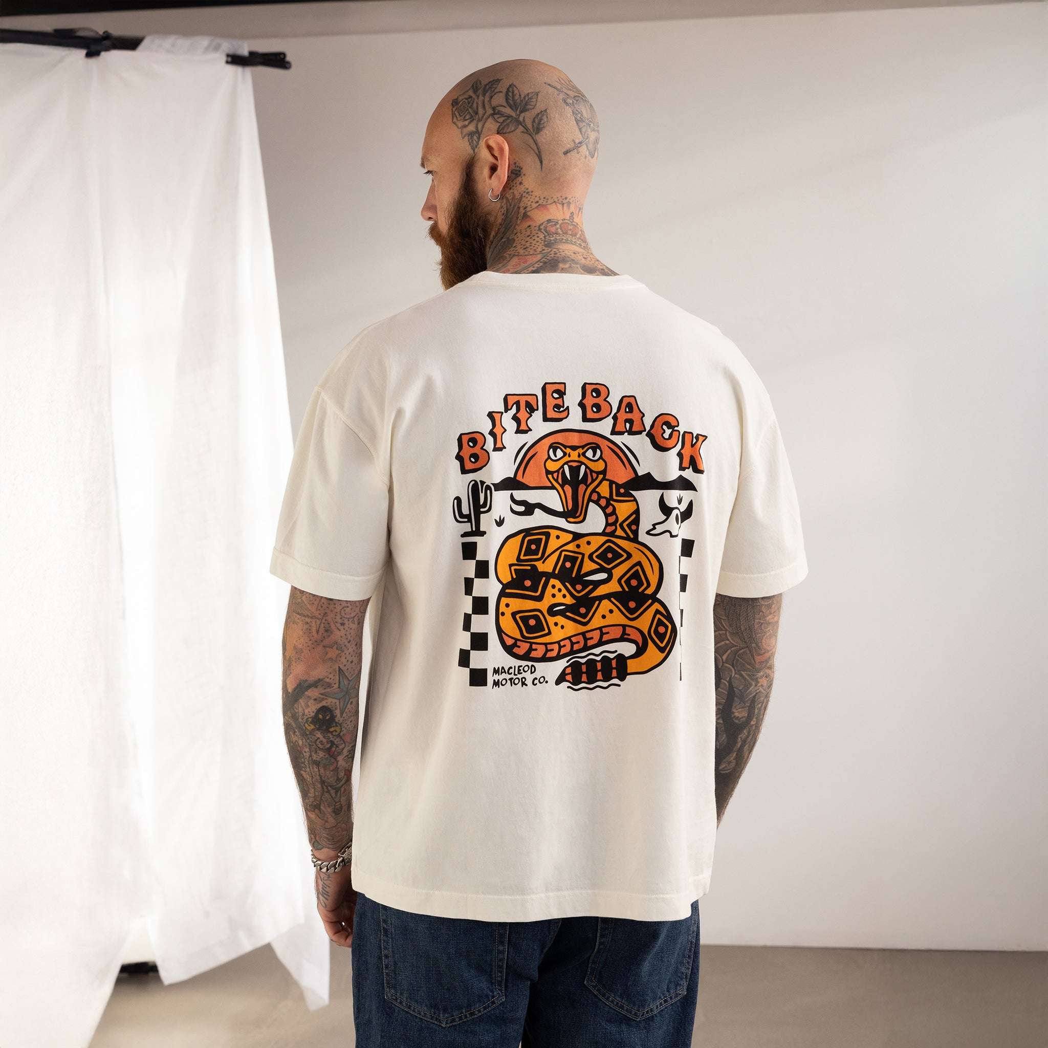 Man wearing a white t-shirt with a snake graphic and text on the back, standing against a plain wall.