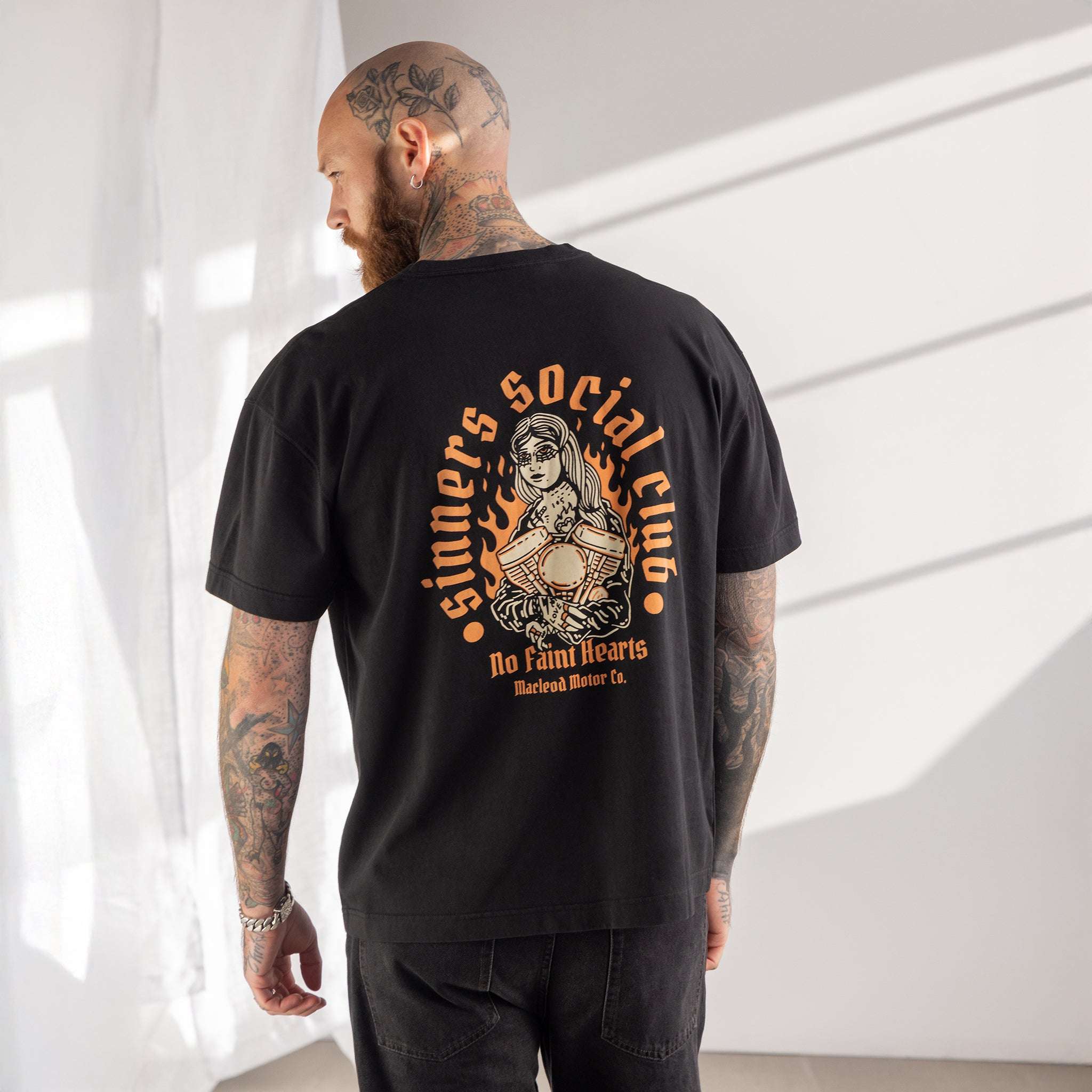 Person wearing a black t-shirt with a graphic design on the back, standing against a white curtain.