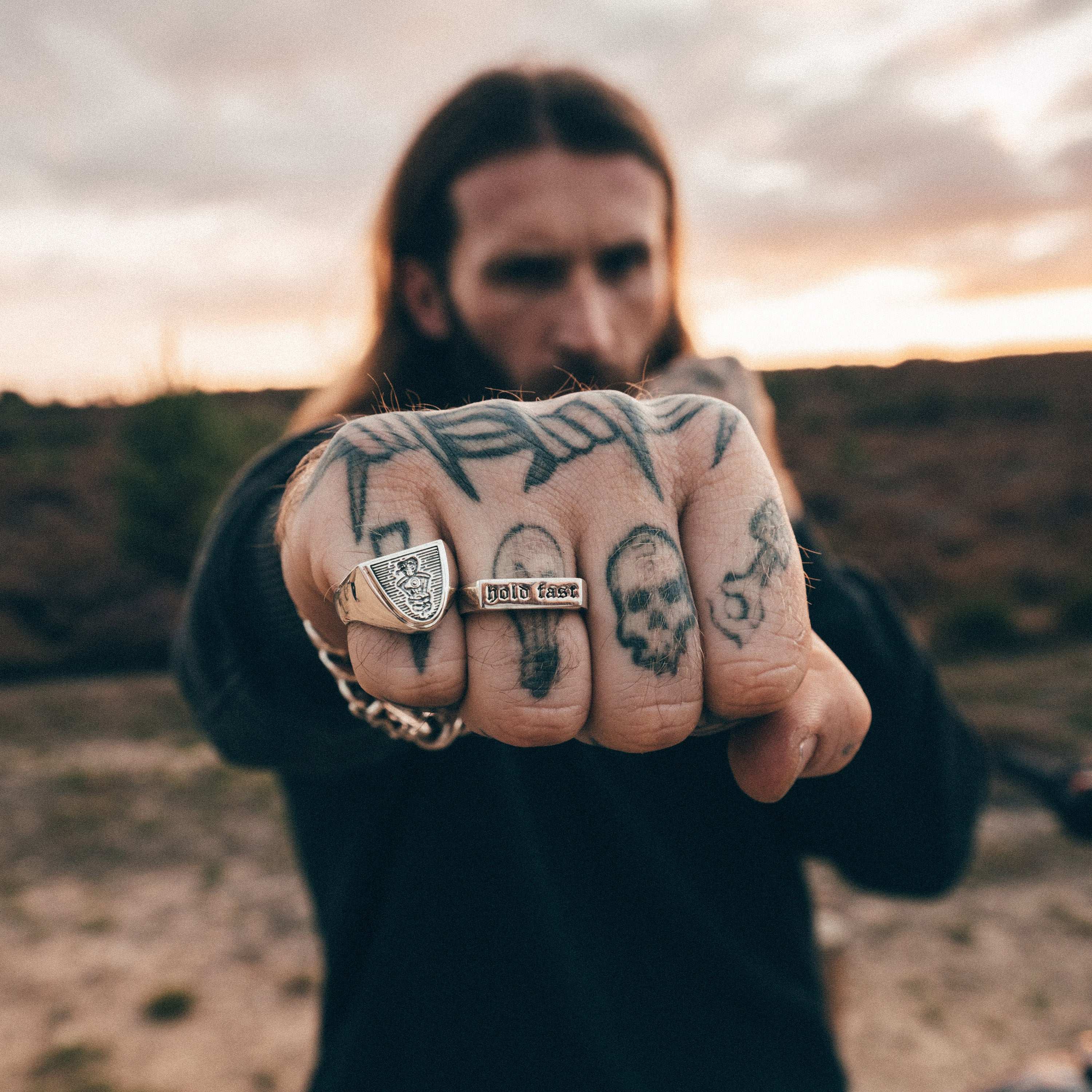 Man with tattoos on his knuckles and a chain in a desert landscape at sunset.