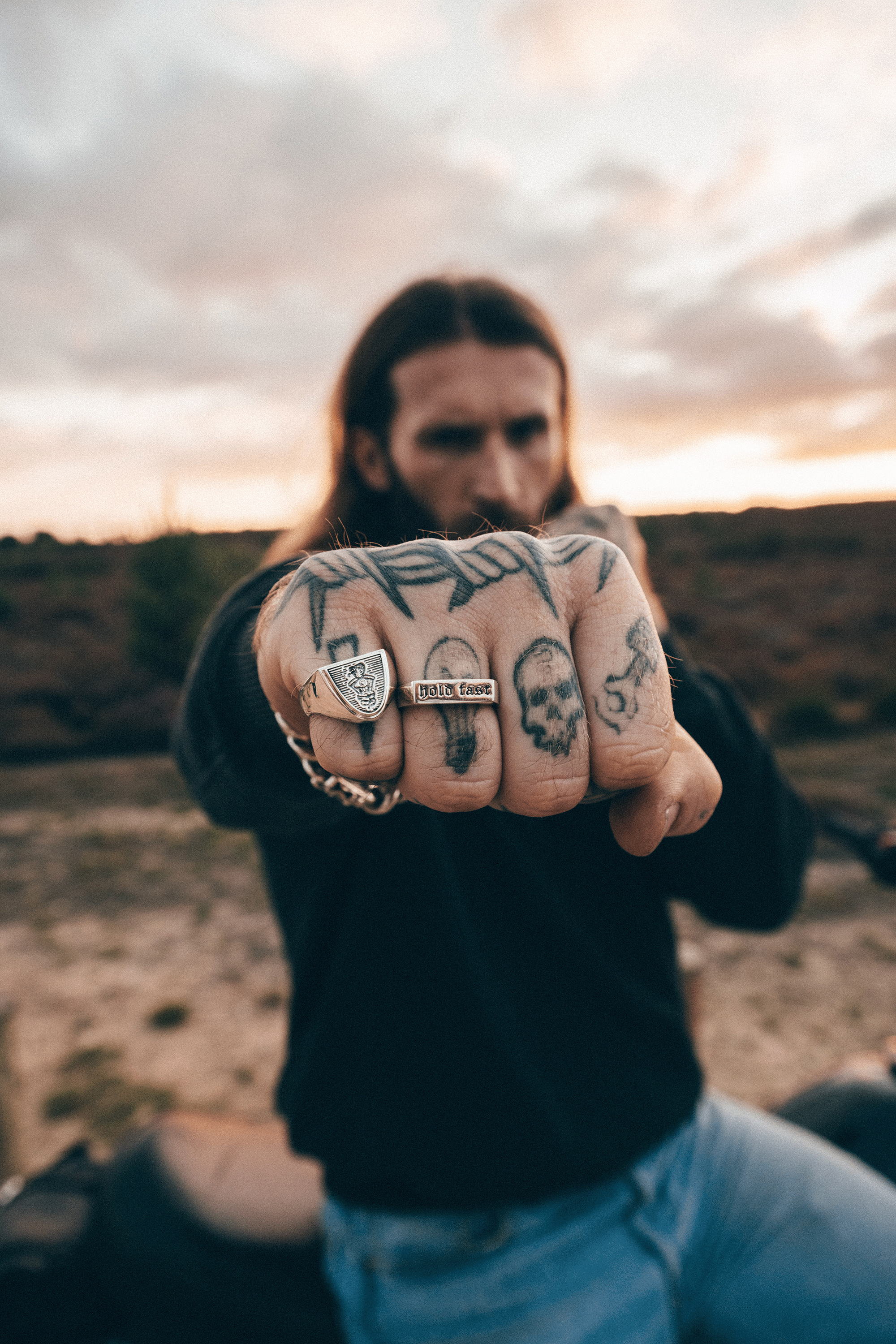 Man with tattoos on his knuckles holding them out towards the camera with a desert landscape in the background.