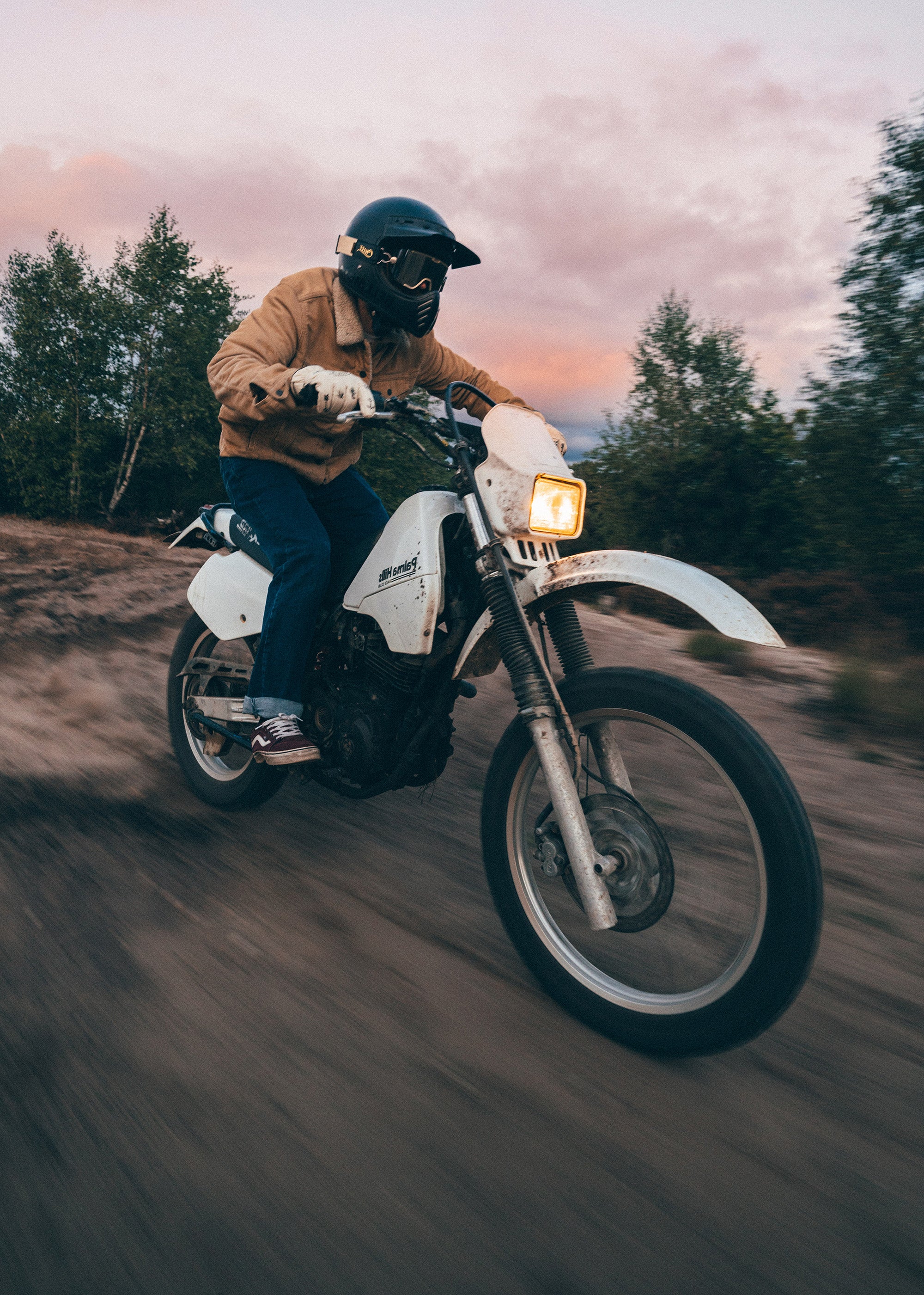 Person riding a motorcycle on a dirt road with trees and a sunset in the background
