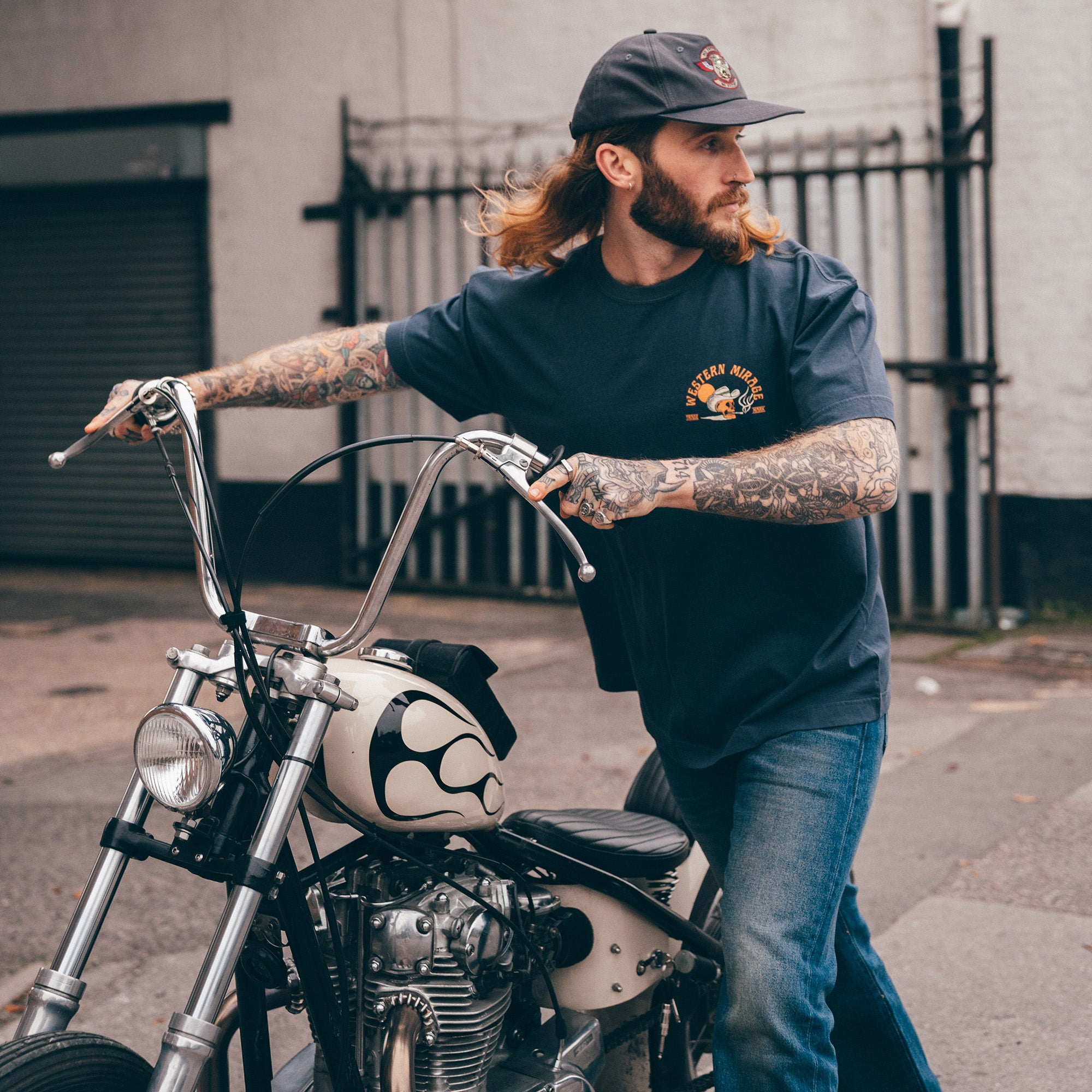 Man with tattoos leaning on a motorcycle wearing a navy blue t-shirt and jeans.